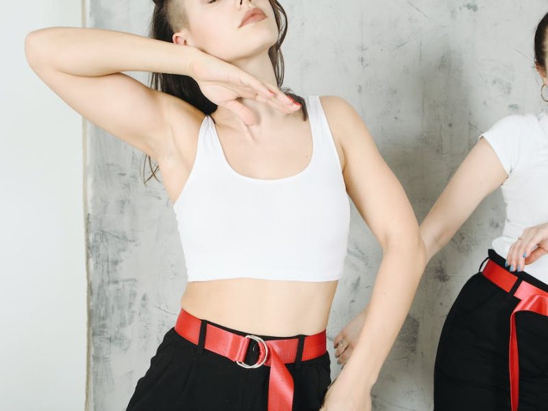 Crop young female dancers in same clothes with red belt and in black pants making dancing movements near gray wall